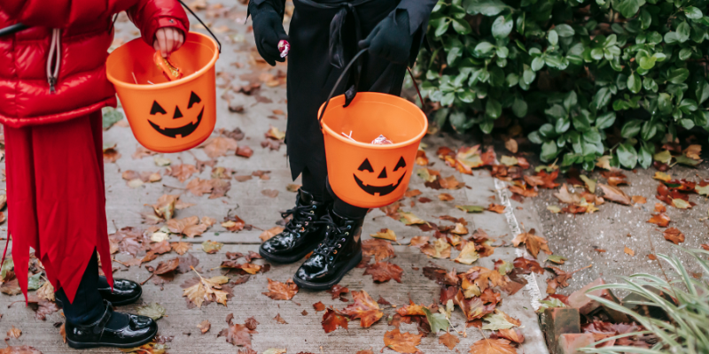 Two children, accompanied by their co-parent, holding pumpkin buckets on a sidewalk as part of their Halloween traditions.