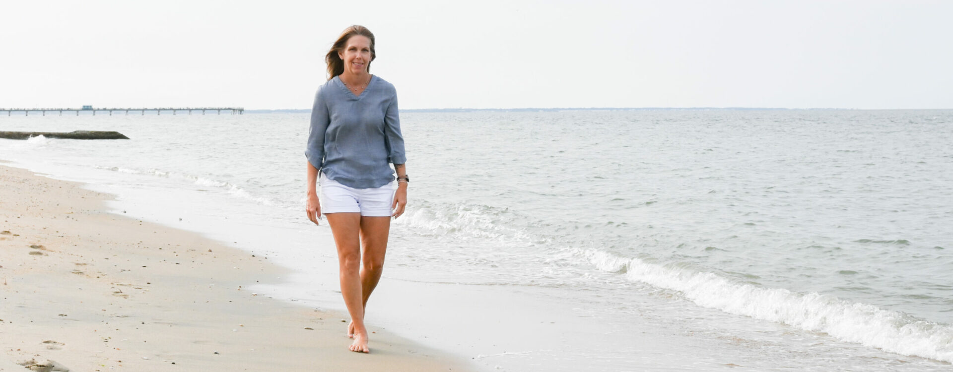 A person walks barefoot along a sandy beach, wearing a blue shirt and white shorts, with gentle waves in the background.